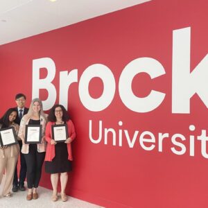 A group of people stand in front of a red wall with the words "Brock University" painted on it.