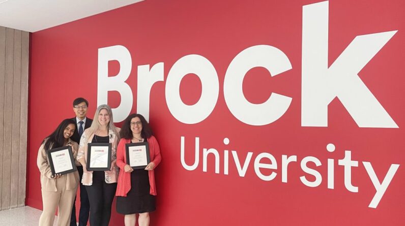 A group of people stand in front of a red wall with the words "Brock University" painted on it.