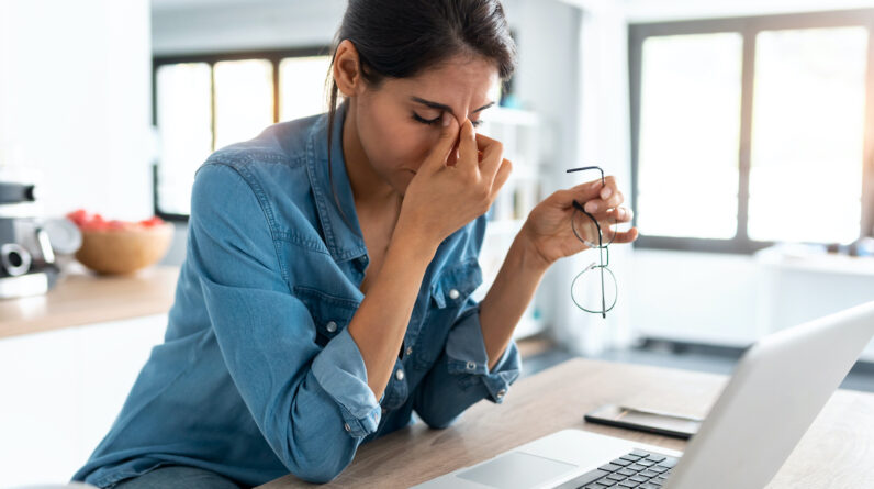 A woman sits at her laptop, pinching the bridge of her nose in frustration.