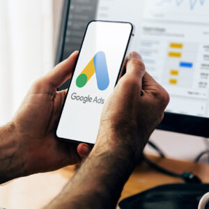 Man using a mobile phone with Google Ads on the screen and in the background a computer showing ad statistics.