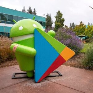 Statue of Android with play store logo amidst plants and grass in garden outside office building at Google campus during sunset.