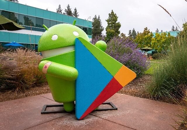 Statue of Android with play store logo amidst plants and grass in garden outside office building at Google campus during sunset.