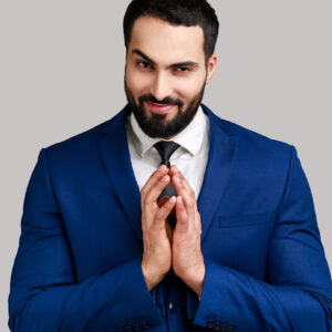 Cunning bearded man clasping hands and planning evil tricky prank or scheming, cheating with sly smile, wearing official style suit. Indoor studio shot isolated on gray background.