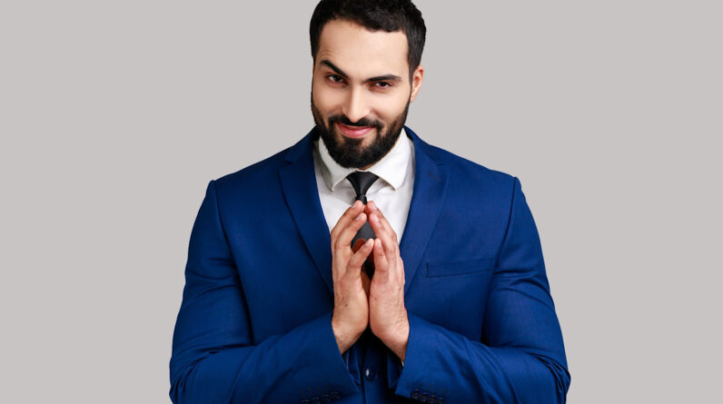 Cunning bearded man clasping hands and planning evil tricky prank or scheming, cheating with sly smile, wearing official style suit. Indoor studio shot isolated on gray background.